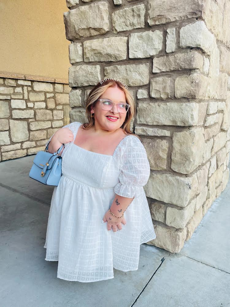 Jonna, a short stature woman standing at 3ft tall wears a white dress and holds a mini blue bag