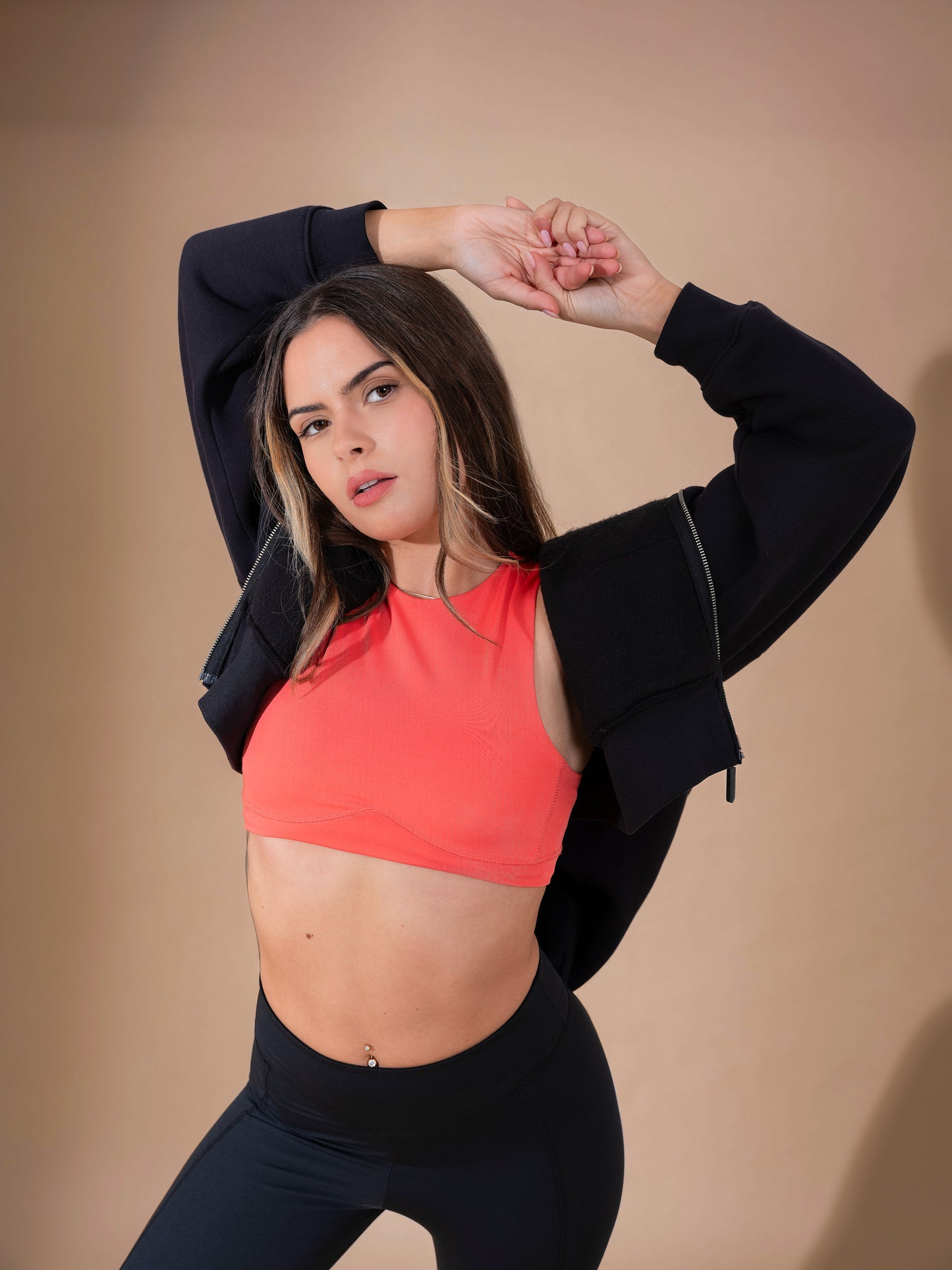A woman in DEWEY's HAZEL Cropped Vest Top (Coral Red, Extra Petite) stretches with arms raised, paired with black leggings and a black jacket against a beige background.