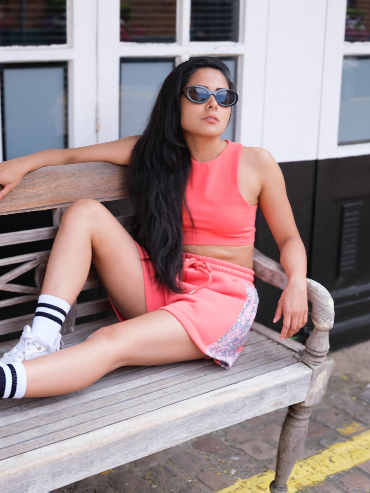 A woman wears the DEWEY HAZEL Cropped Vest Top in Coral Red (Extra Petite) with shorts, sunglasses, and sneakers as she sits relaxed on a wooden bench outdoors—showcasing an easy athleisure style.