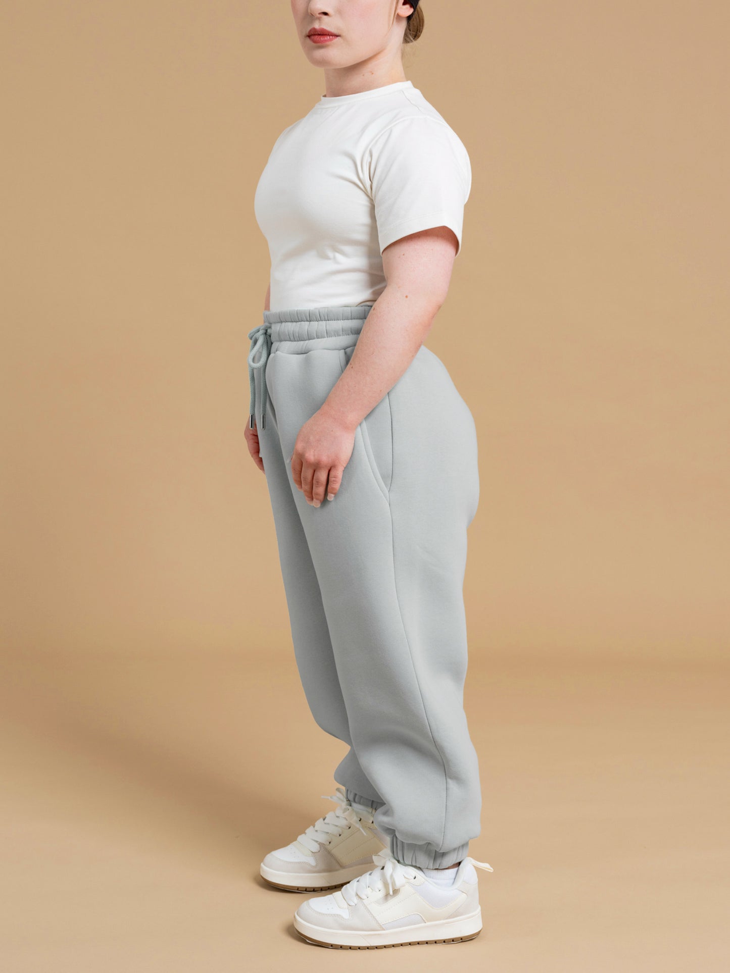 A person stands sideways against a beige background, wearing the DEWEY ALEX Slim Fit Short Sleeve T-Shirt Tee in White (Extra Petite), light blue jogger sweatpants, and white sneakers.