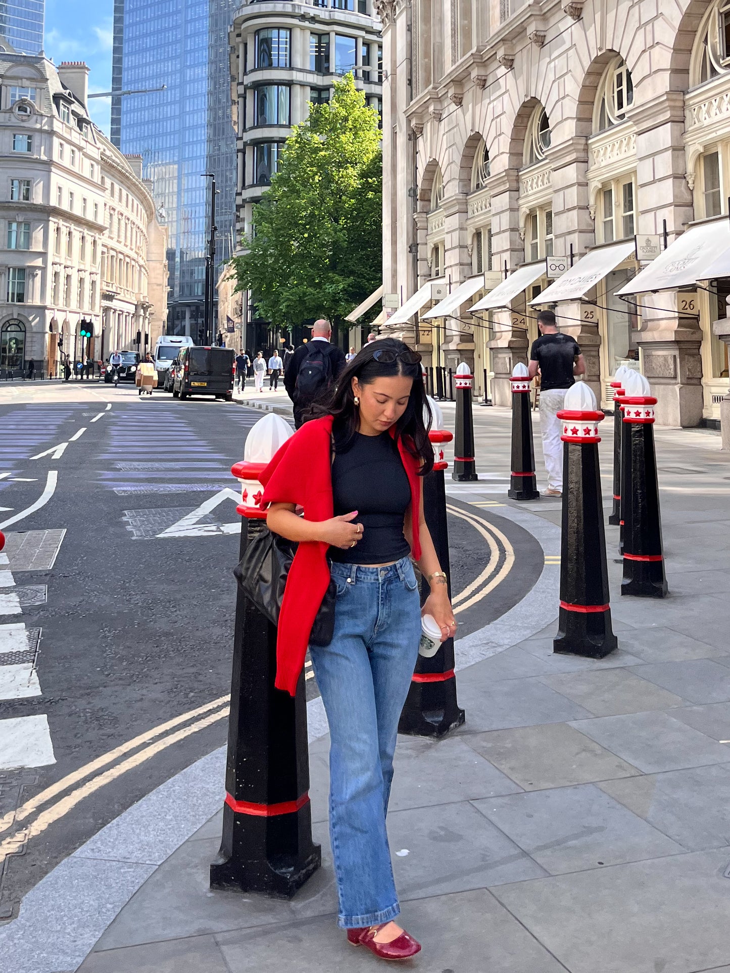 A woman in a red cardigan, blue jeans, and red shoes walks down a city street holding a cup and carrying a black bag. She wears the DEWEY SUMMER Racerback Vest Top (Black, Extra Petite) against a backdrop of historic buildings and skyscrapers.