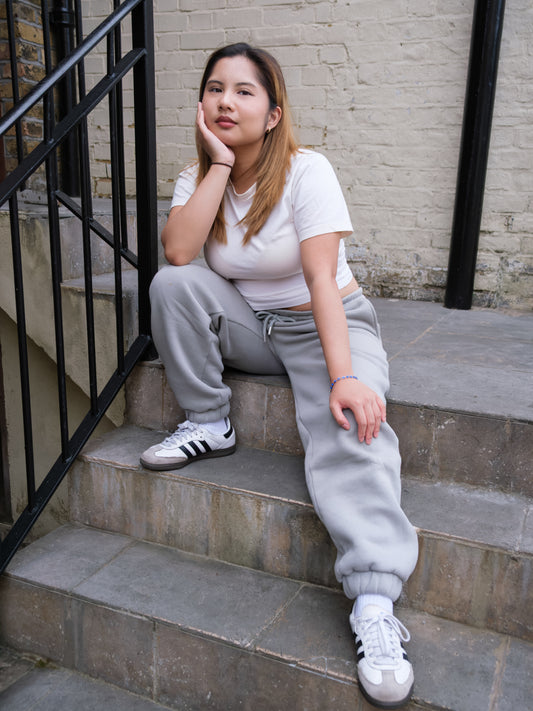 A young woman with straight, light brown hair sits on outdoor steps, relaxed and thoughtful. She wears a white t-shirt, DEWEY RILEY Sweatpants Joggers in grey (Extra Petite), and white Adidas sneakers—perfect loungewear for petites.