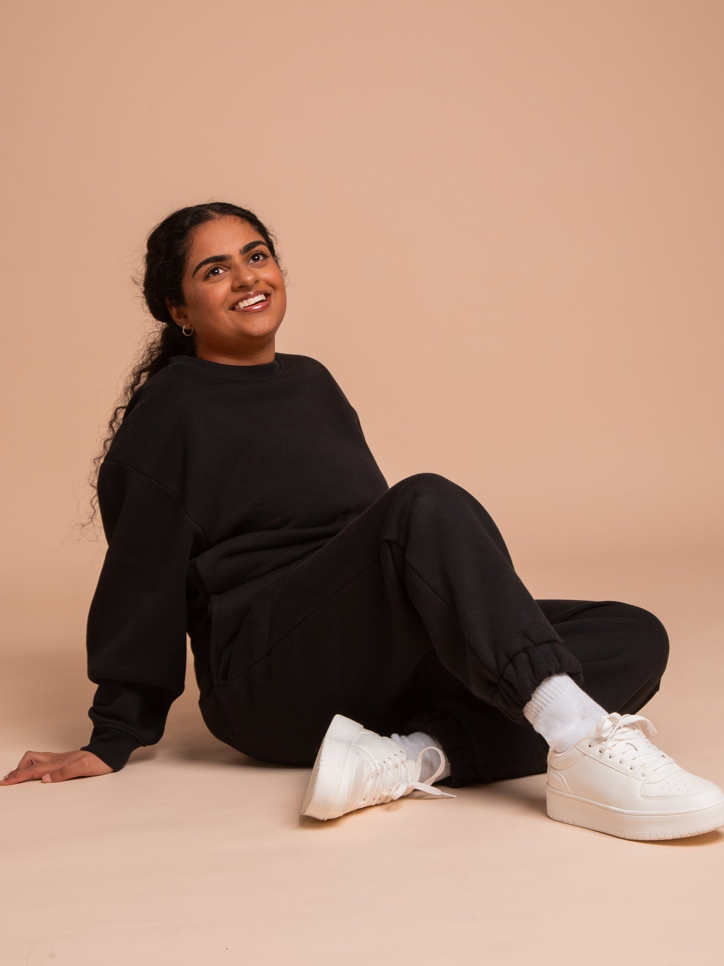 A woman with long dark hair smiles and looks up as she sits on the floor against a beige background, wearing a relaxed fit DEWEY BAILEY Crewneck Sweatshirt Jumper in black (Extra Petite), matching joggers, white socks, and white sneakers.