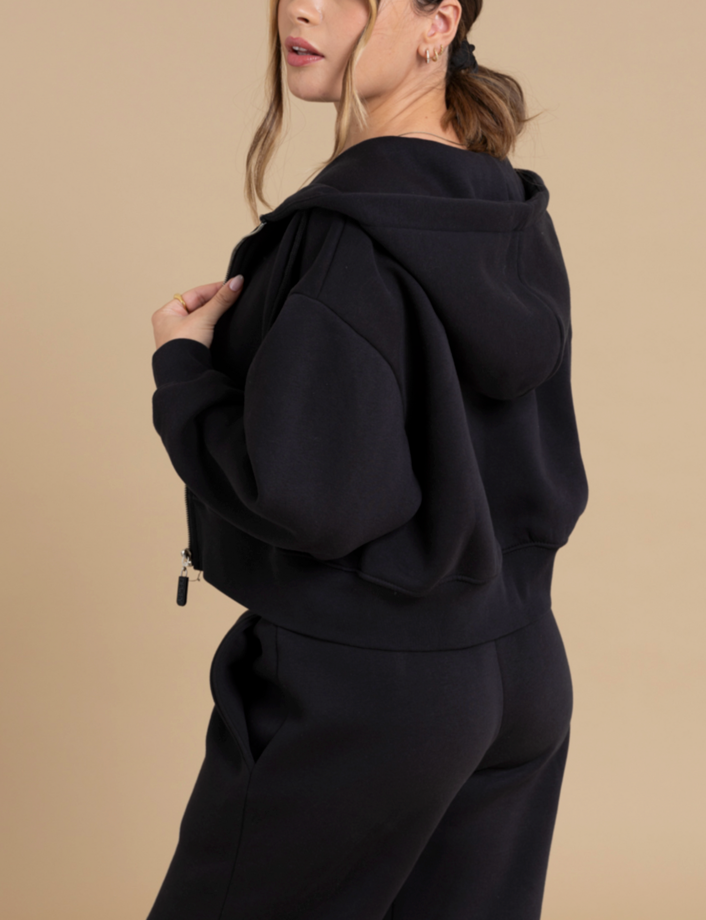 A woman in a DEWEY TOBY Full Zip-Up Hoodie (Black, Extra Petite) and matching sweatpants stands against a beige background, looking over her shoulder with one hand on her hoodie’s collar.