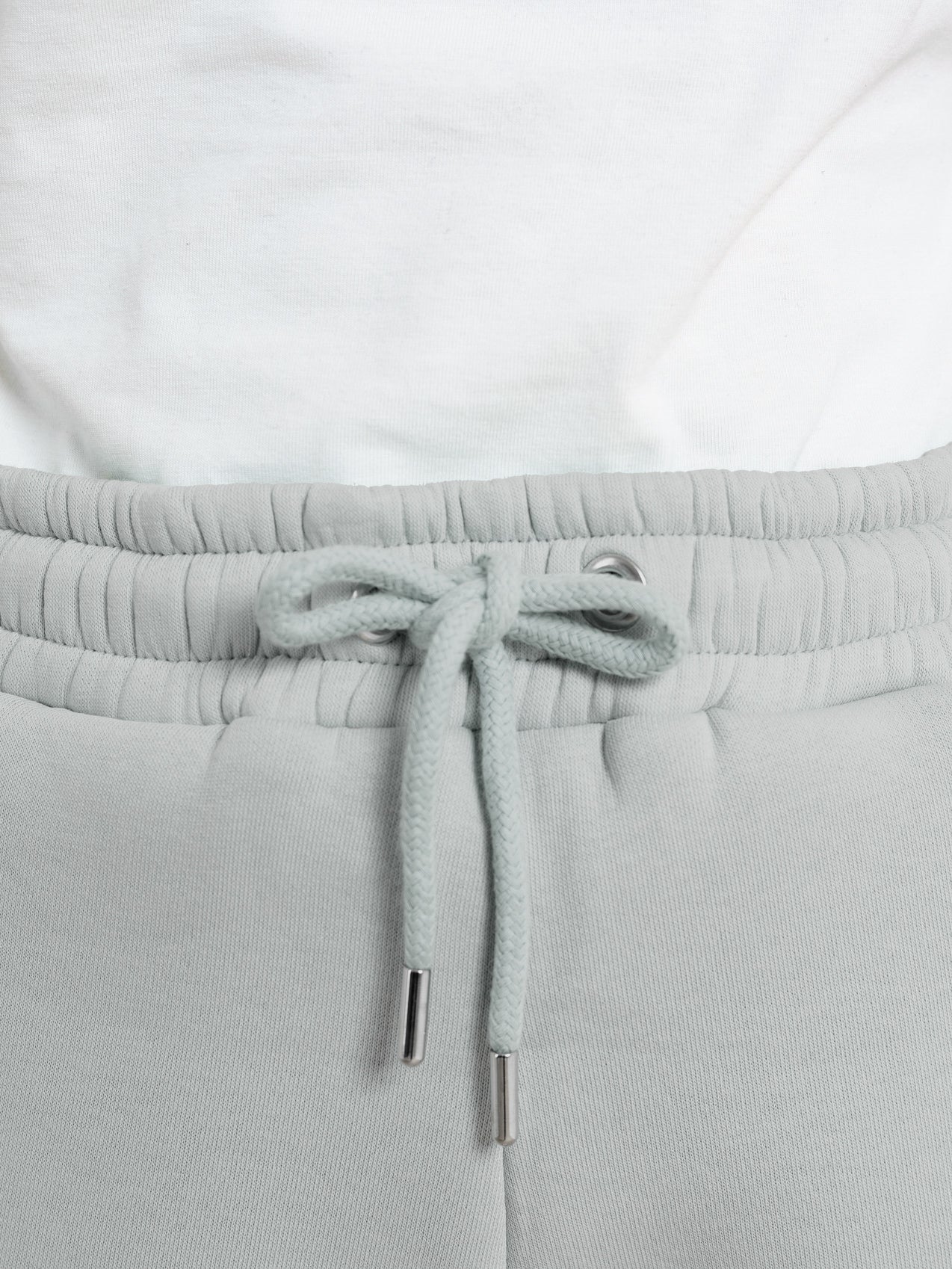 Close-up of elevated loungewear: a person wears DEWEY's RILEY Sweat Shorts in grey (Extra Petite) with a drawstring tied in a bow, paired with a white shirt tucked at the waist.