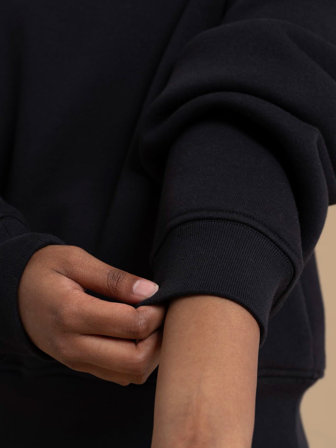 A person adjusts the sleeve of a relaxed fit BAILEY Crewneck Sweatshirt Jumper by DEWEY in black (Extra Petite), pulling the cuff up their forearm with their other hand against a neutral background, focusing on the sleeve and hand.