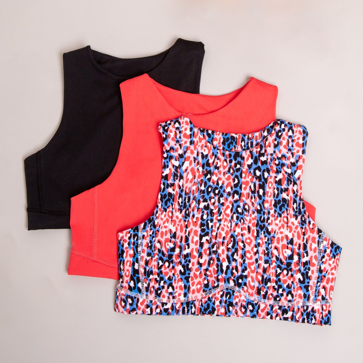 Three cropped tank tops are laid flat: one solid black, one solid red, and a DEWEY HAZEL Cropped Vest Top (Printed, Extra Petite) in a red, blue, and white leopard print. The athleisurewear pieces overlap on a neutral background.