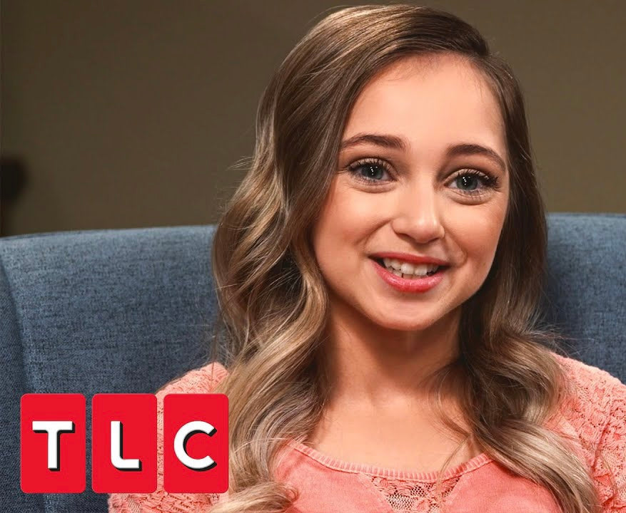 Shauna Rae is a 23 year old, light olive-skinned woman with straight dirty blonde-brown hair and blue eyes. She is 3’10” and wears a salmon pink top. In the bottom left corner is the TLC logo from her show
