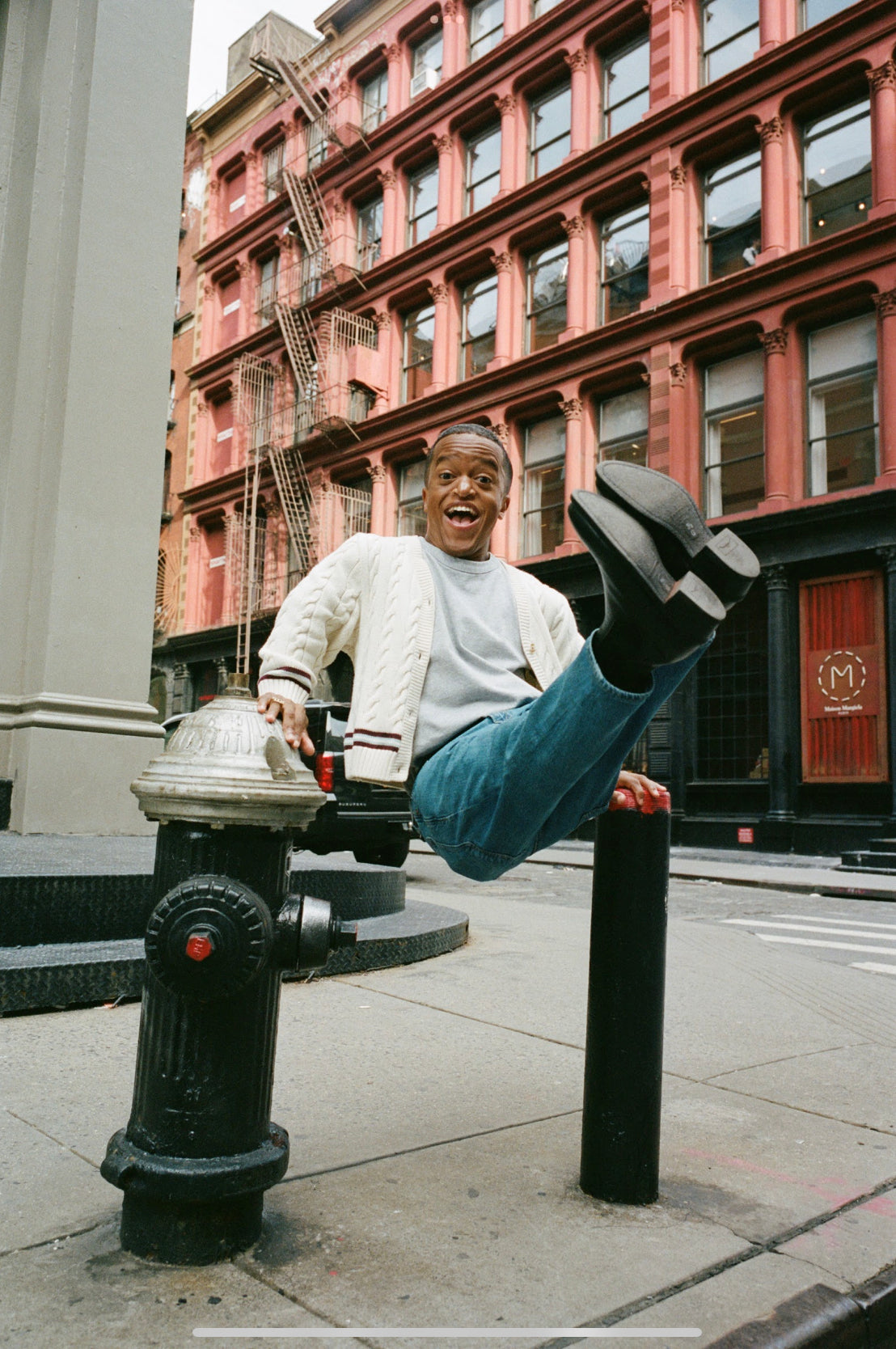 A 31 year old, straight African American man with short hair and dark brown eyes, standing 4'6" tall. He wears blue colour trousers, black boots, a white tshirt with a cream cardigan. He is elevated off two bollards, seemingly in the USA
