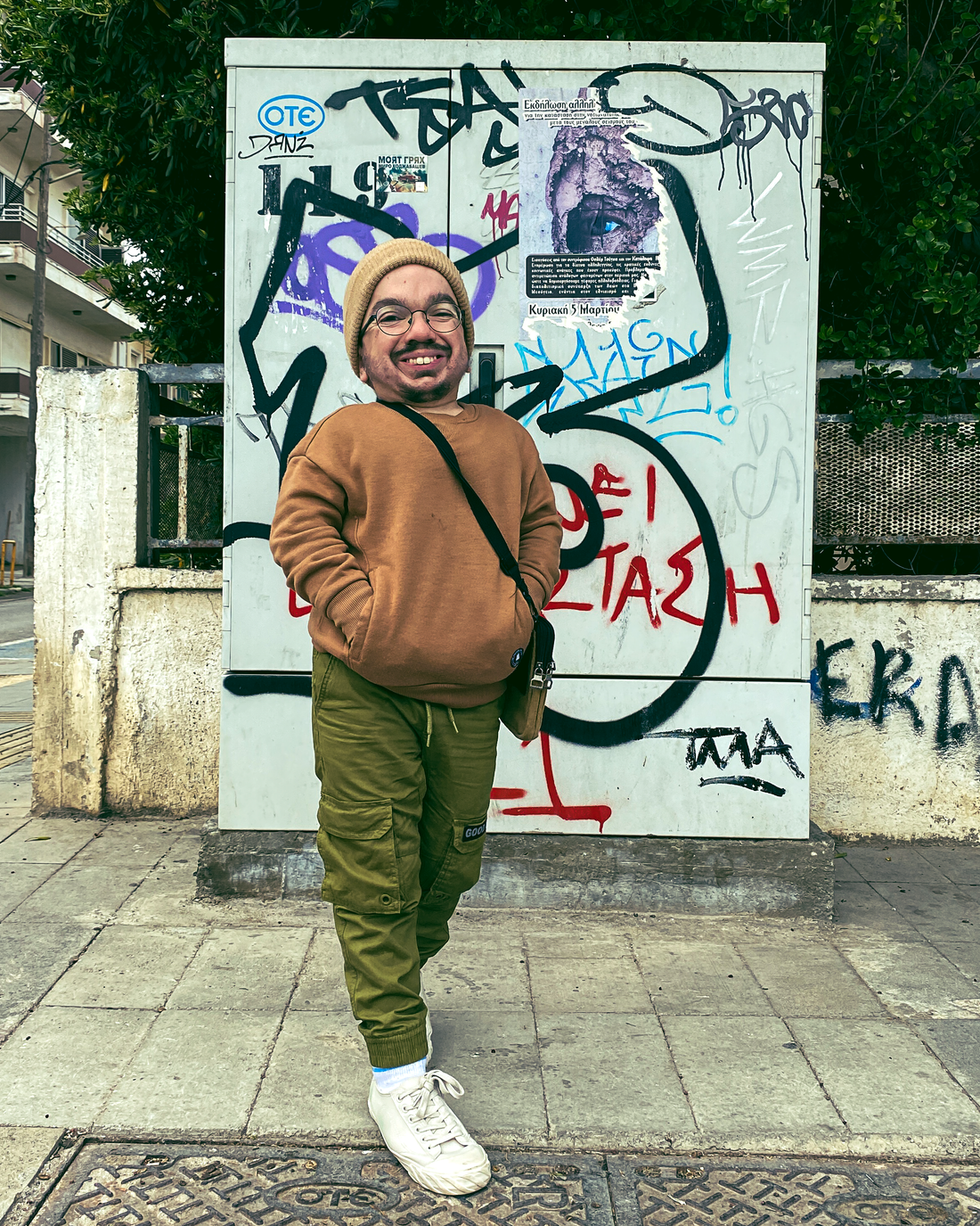 Oscar, a short stature petite male with fair skin and brown facial hair and glasses wears a beige/brown jumper with green cargo trousers and white trainers with a crossbody bag. Behind him is graffiti 