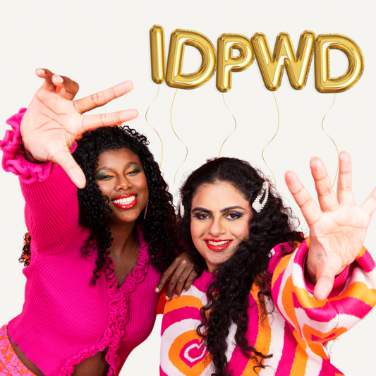 Two women wearing vibrant pink tops reach their hands to the camera, above them is gold balloons spelling out ‘IDPWD’
