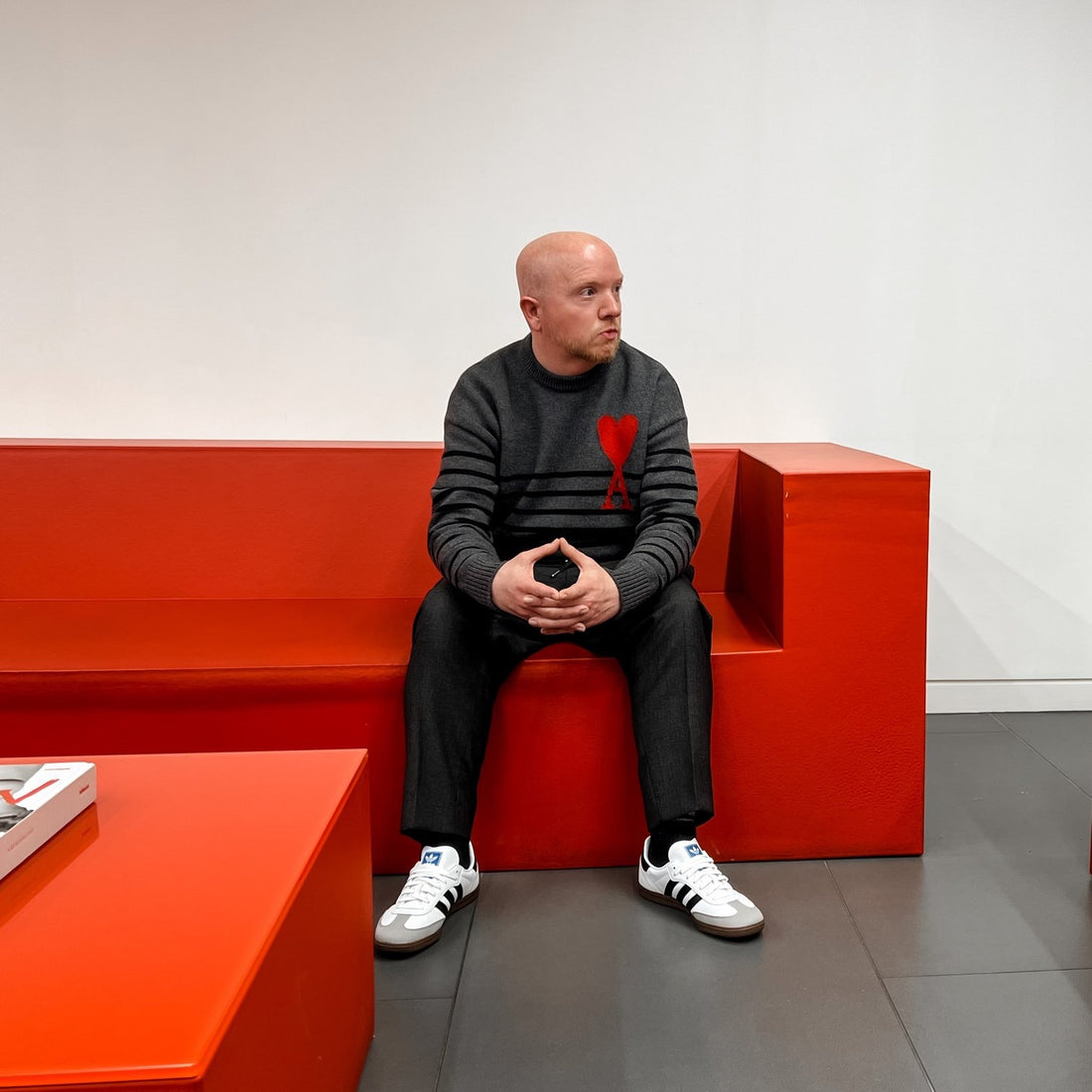Kris is a bald fair-skinned man. He is sat on a red blocky sofa, wearing a grey striped jumper, with black trousers and white trainers