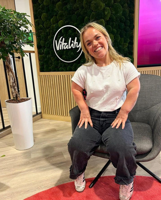 Ellie Simmonds who is 28year old woman with dwarfism who has blonde wavy hair with blue eyes and a fair complexion. She is sat on a chair with a big cheesy grin on her face. She wears a white tshirt with black jeans.