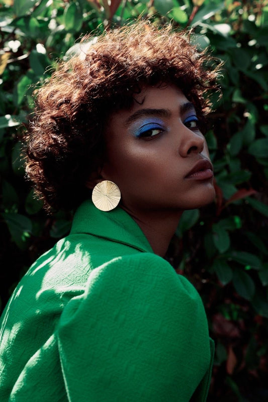 A woman with a deep, rich skin tone and curly Afro hair looks over her shoulder towards the camera. She wears blue eyeshadow, and a green blazer