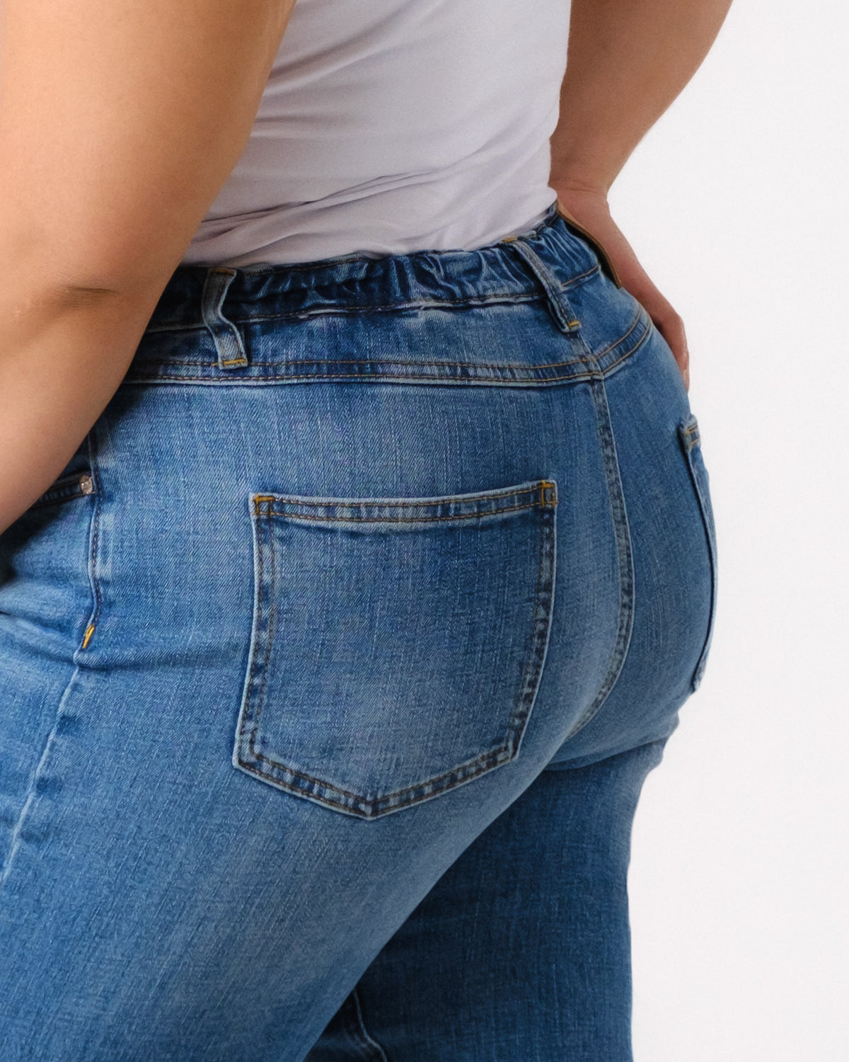 A person wearing a white top and blue denim jeans is shown from the waist down, with focus on the back pocket and side seams of the jeans.
