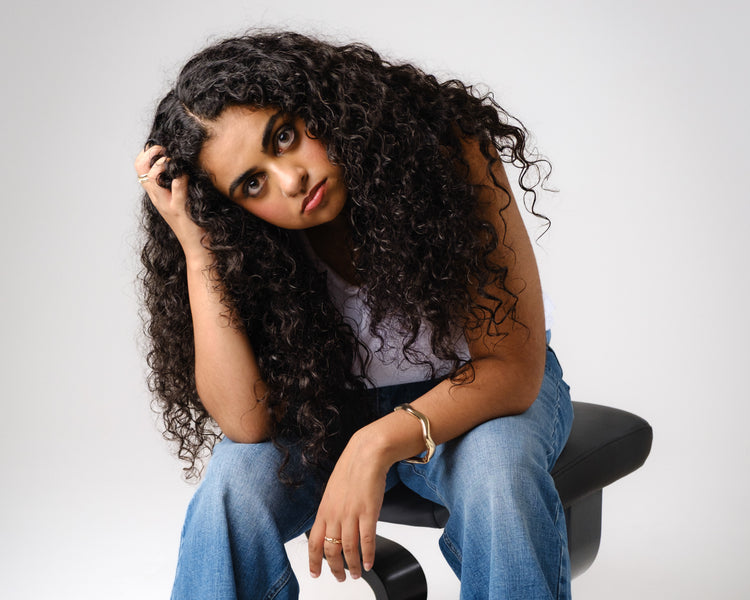 A person wearing a white top and black jeans stands with hands on hips, showing long white nails, chunky rings, pearl bracelets, and gold bangles against a neutral background.
