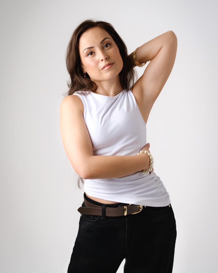 A woman with medium-length dark hair wearing a white sleeveless top and black pants stands against a beige background, resting her chin on her hand and looking confidently at the camera.