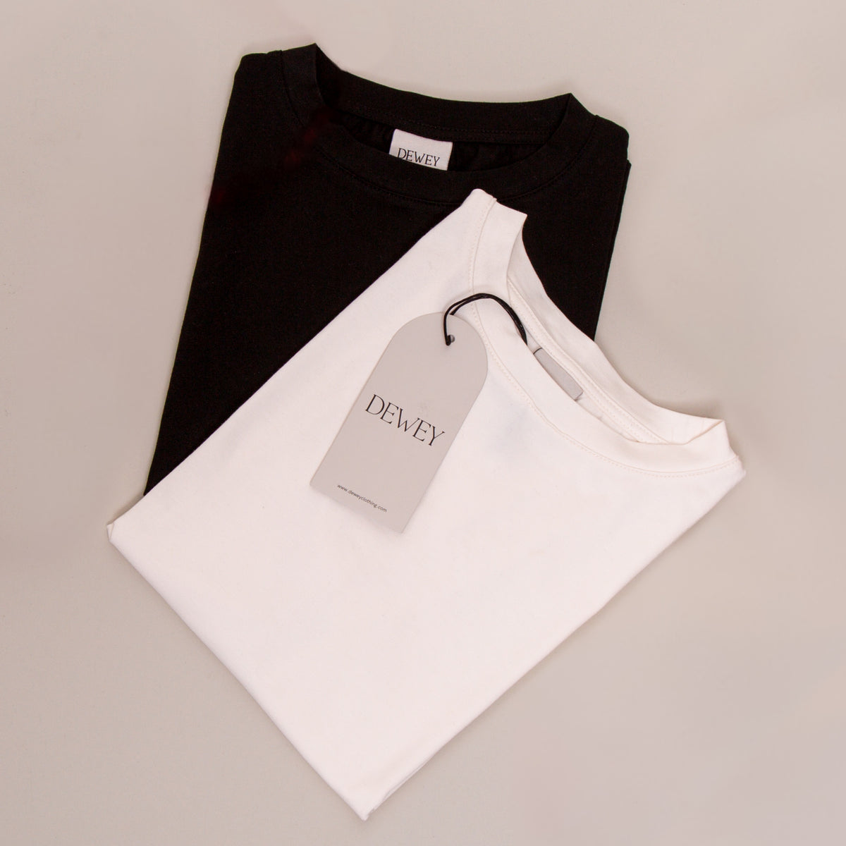 Two folded t-shirts, one black and one white, are stacked on a light surface. The white t-shirt is on top and has a DEWEY brand tag attached to its collar.