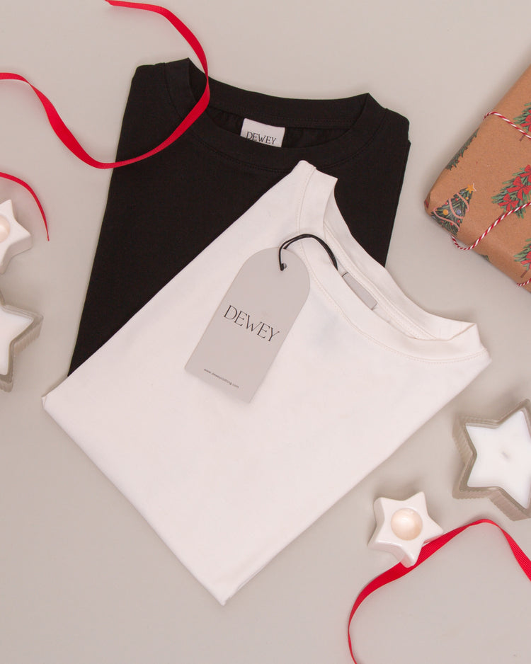 Two folded t-shirts, one black and one white with a “DEWEY” tag, lie on a light surface surrounded by red ribbon, gift-wrapped present, and star-shaped candles.