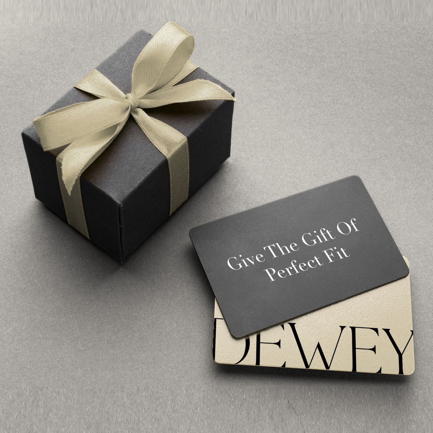 A black gift box with a cream ribbon sits next to two overlapping gift cards. The top card reads "Give The Gift Of Perfect Fit" in white text on a black background.