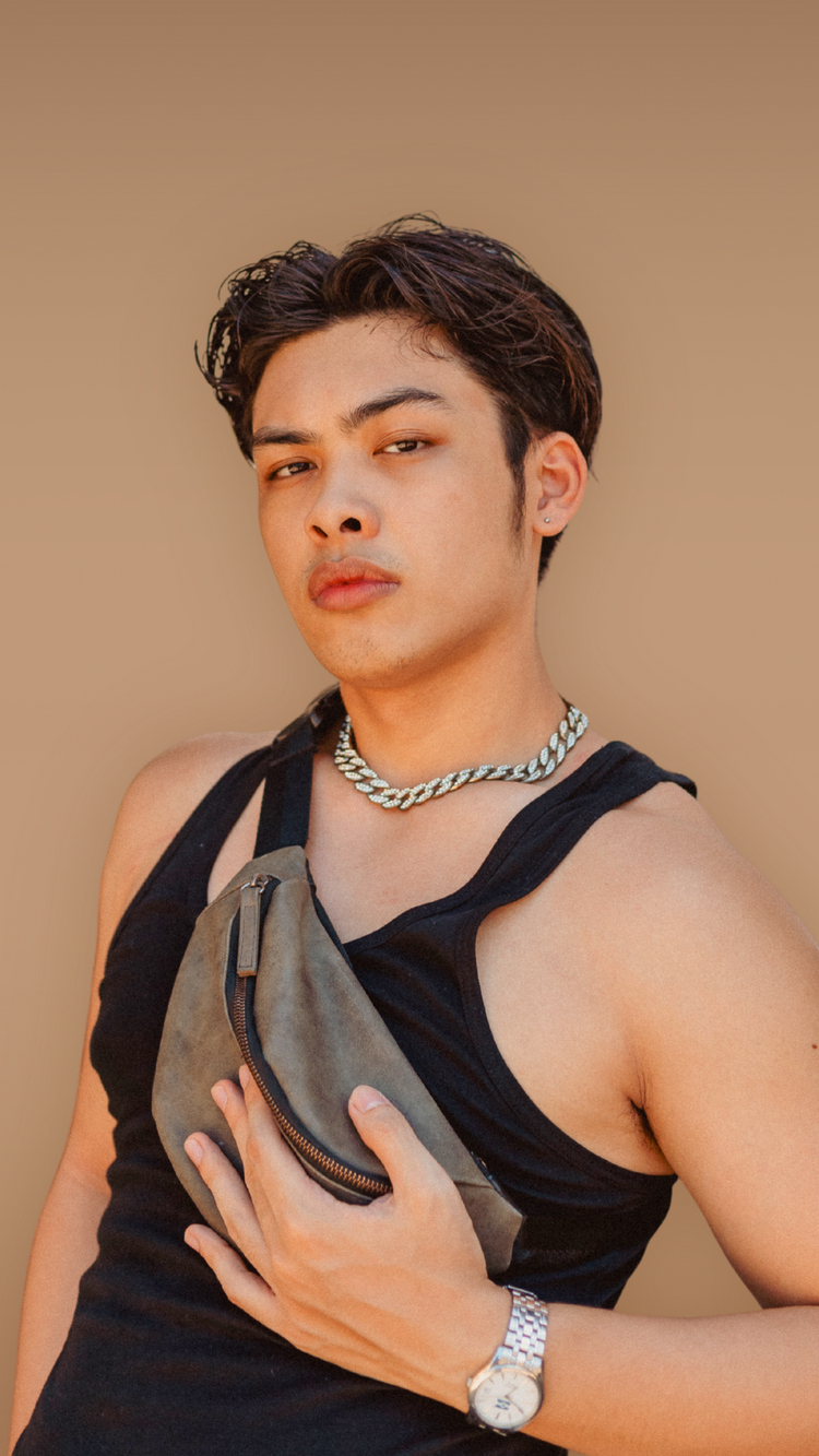A person wearing a black tank top and silver chain necklace, posing against a brown background. They have a gray bag slung across their chest and a wristwatch on their left hand.