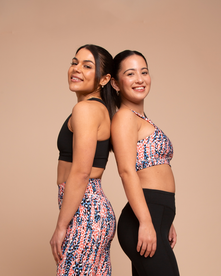 Two women in activewear stand back-to-back, smiling. One wears a black sports bra and patterned leggings, the other a patterned sports bra and black leggings. The background is a warm beige color.