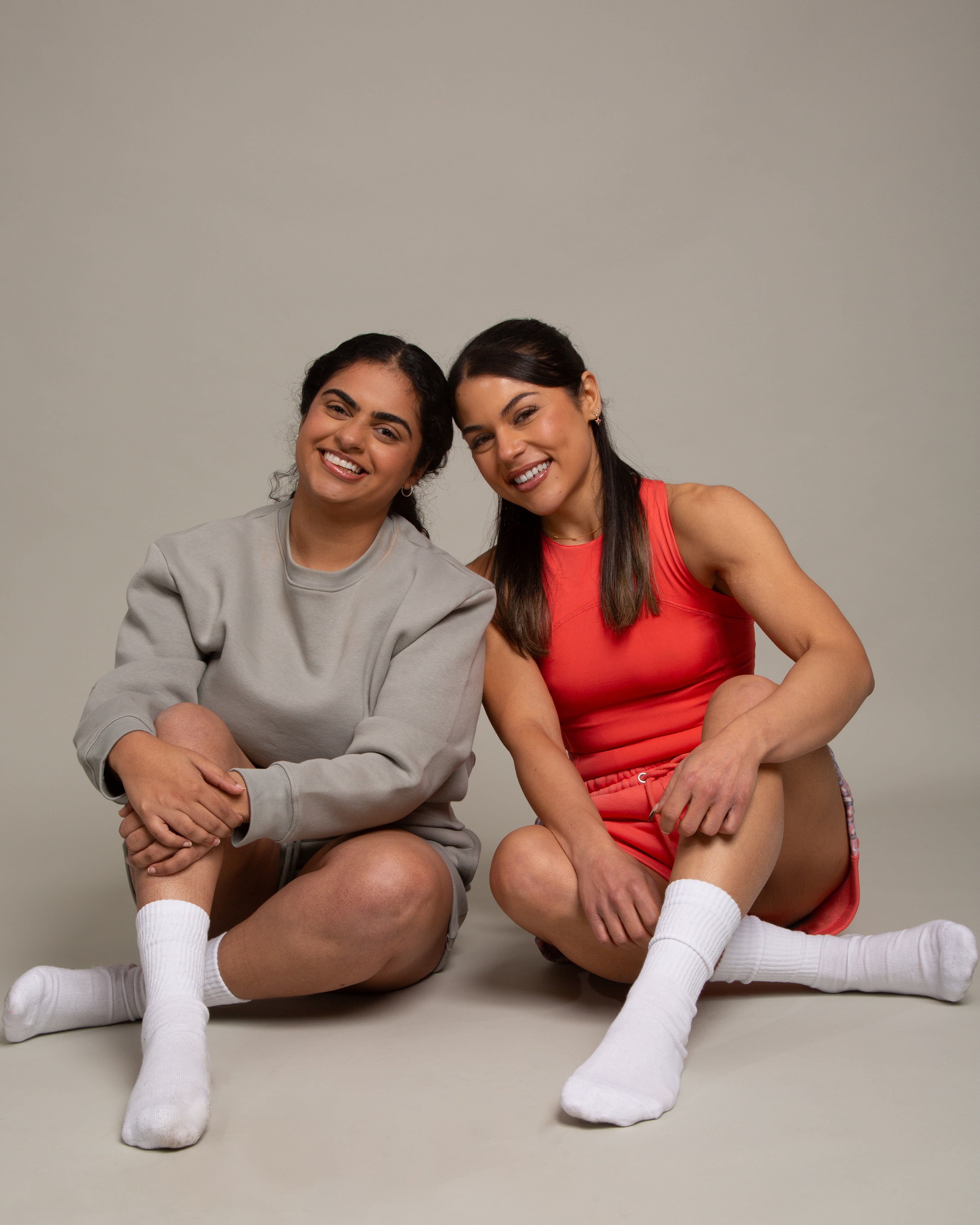 Two women with dark hair and white socks sit on the floor smiling. One wears a grey sweatshirt and shorts; the other wears the DEWEY SUMMER Racerback Vest Top in Coral Red (Extra Petite). The plain backdrop highlights their outfits.
