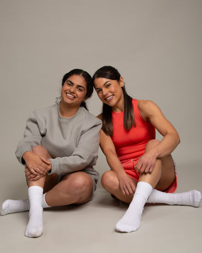 Two women with dark hair and white socks sit on the floor smiling. One wears a grey sweatshirt and shorts; the other wears the DEWEY SUMMER Racerback Vest Top in Coral Red (Extra Petite). The plain backdrop highlights their outfits.