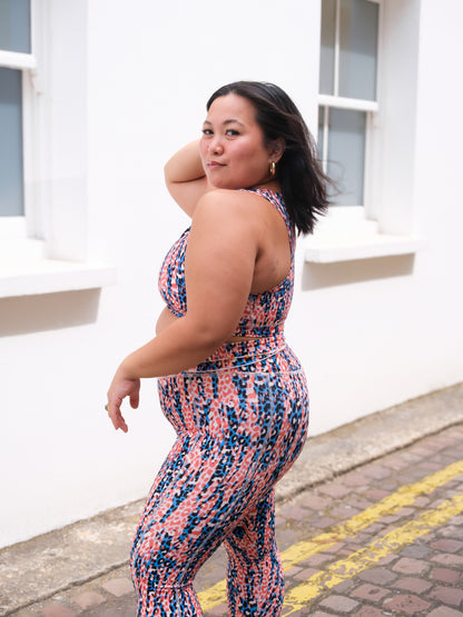 A woman wearing DEWEY's IRIS High Waist Leggings (Printed, Extra Petite) glances over her shoulder while standing on a sidewalk by a white building with large windows.