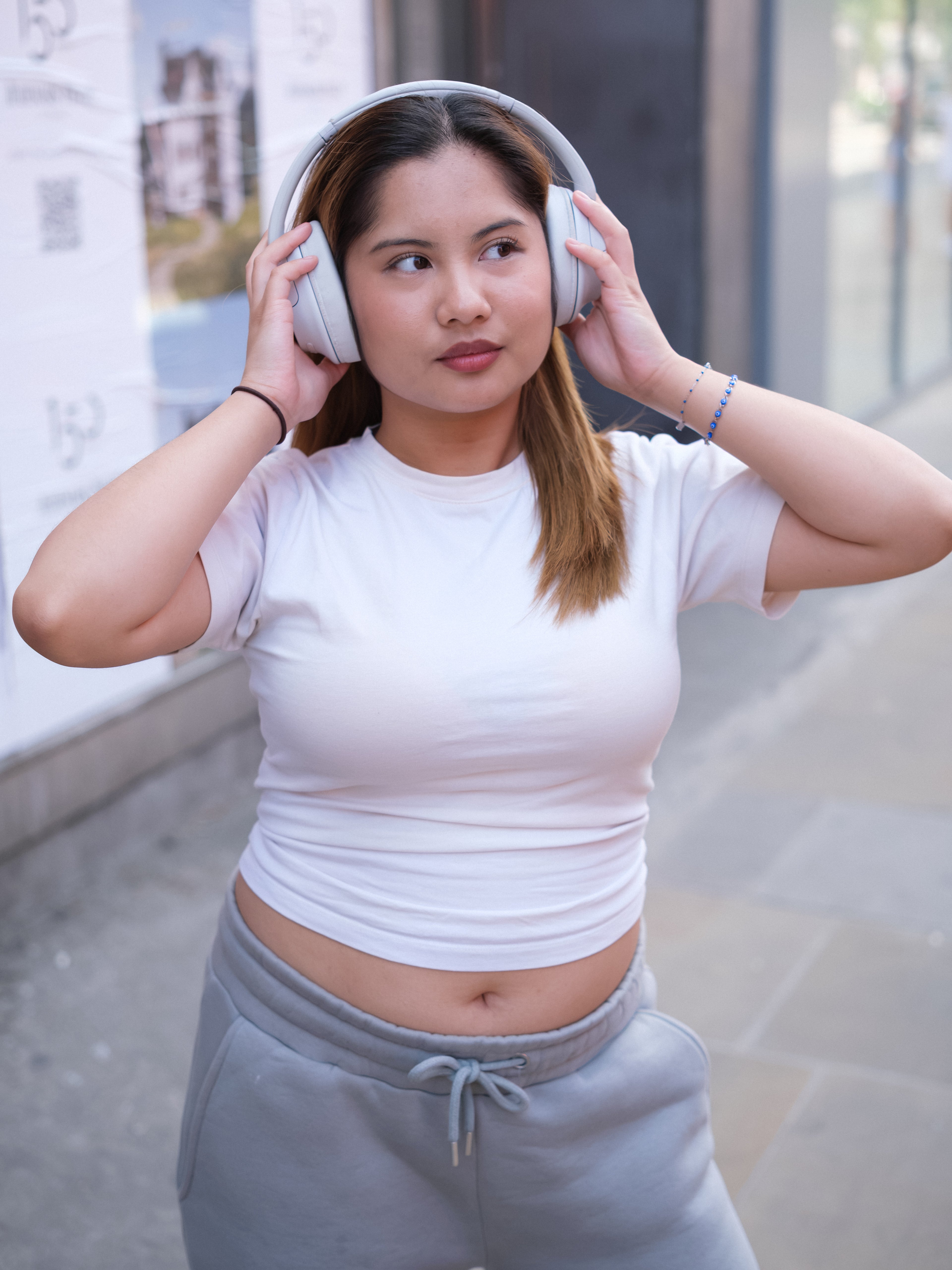 A young woman with light brown hair stands on a city sidewalk in a DEWEY ALEX Slim Fit Short Sleeve T-Shirt Tee (White, Extra Petite) and gray sweatpants, holding over-ear headphones and looking to the side.