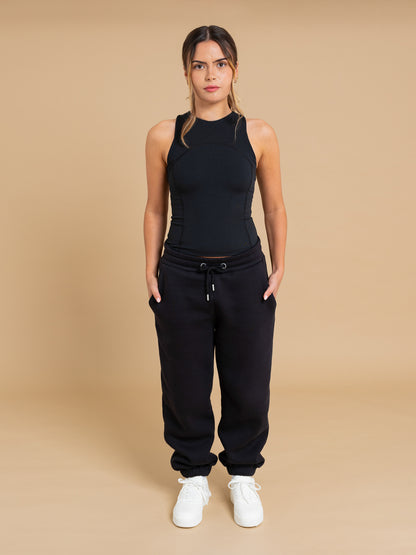 A woman with her hair tied back stands against a beige background wearing the DEWEY SUMMER Racerback Vest Top in black (Extra Petite), paired with black jogger pants with pockets and white sneakers, her hands resting in her pockets.