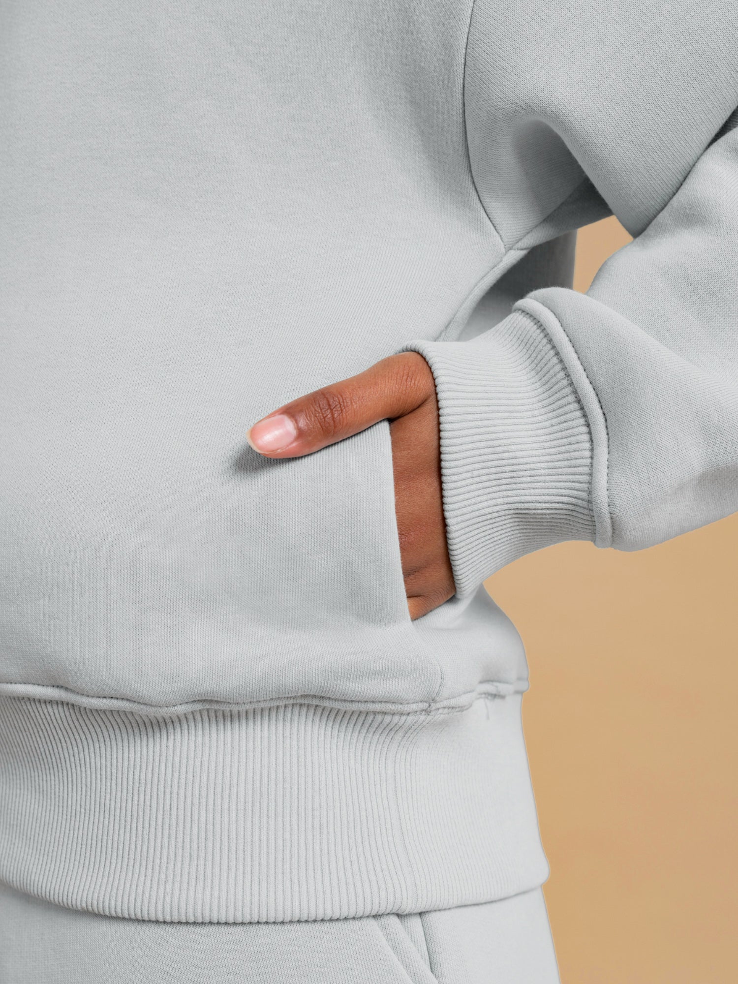 A close-up of a person wearing the DEWEY BAILEY Crewneck Sweatshirt Jumper in Grey (Petite & Extra Petite) with their right hand partially tucked into the front pocket, set against a plain beige background.