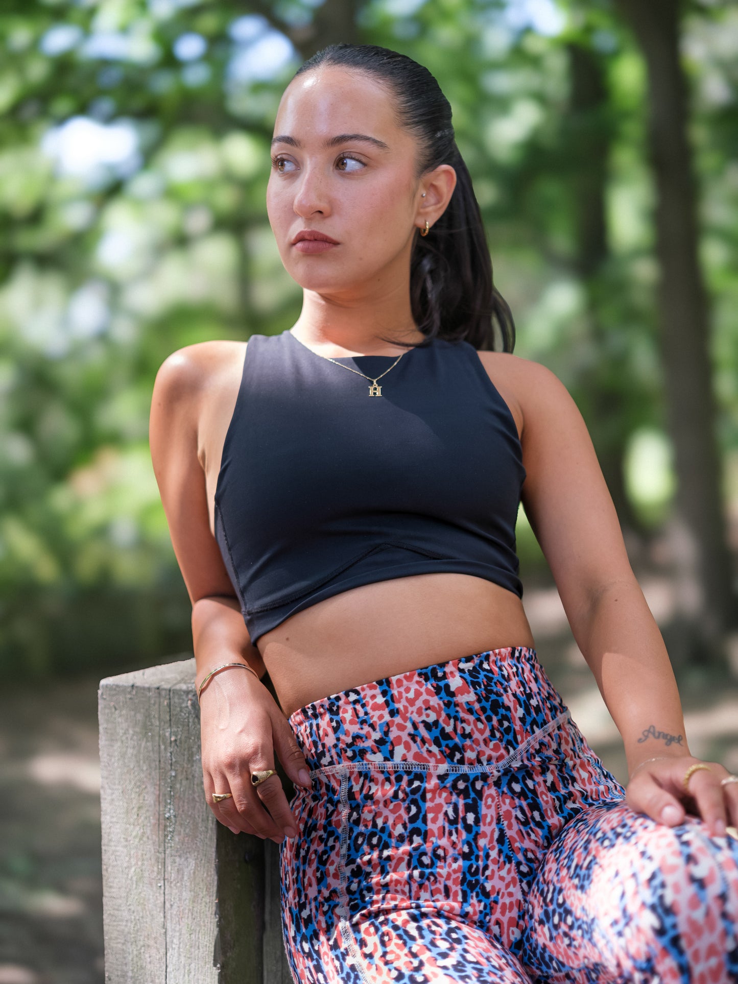 A woman with long dark hair in a ponytail sits thoughtfully on a wooden bench outdoors, wearing a black sleeveless top and DEWEY IRIS High Waist Leggings (Printed, Extra Petite). Blurred trees appear in the background.