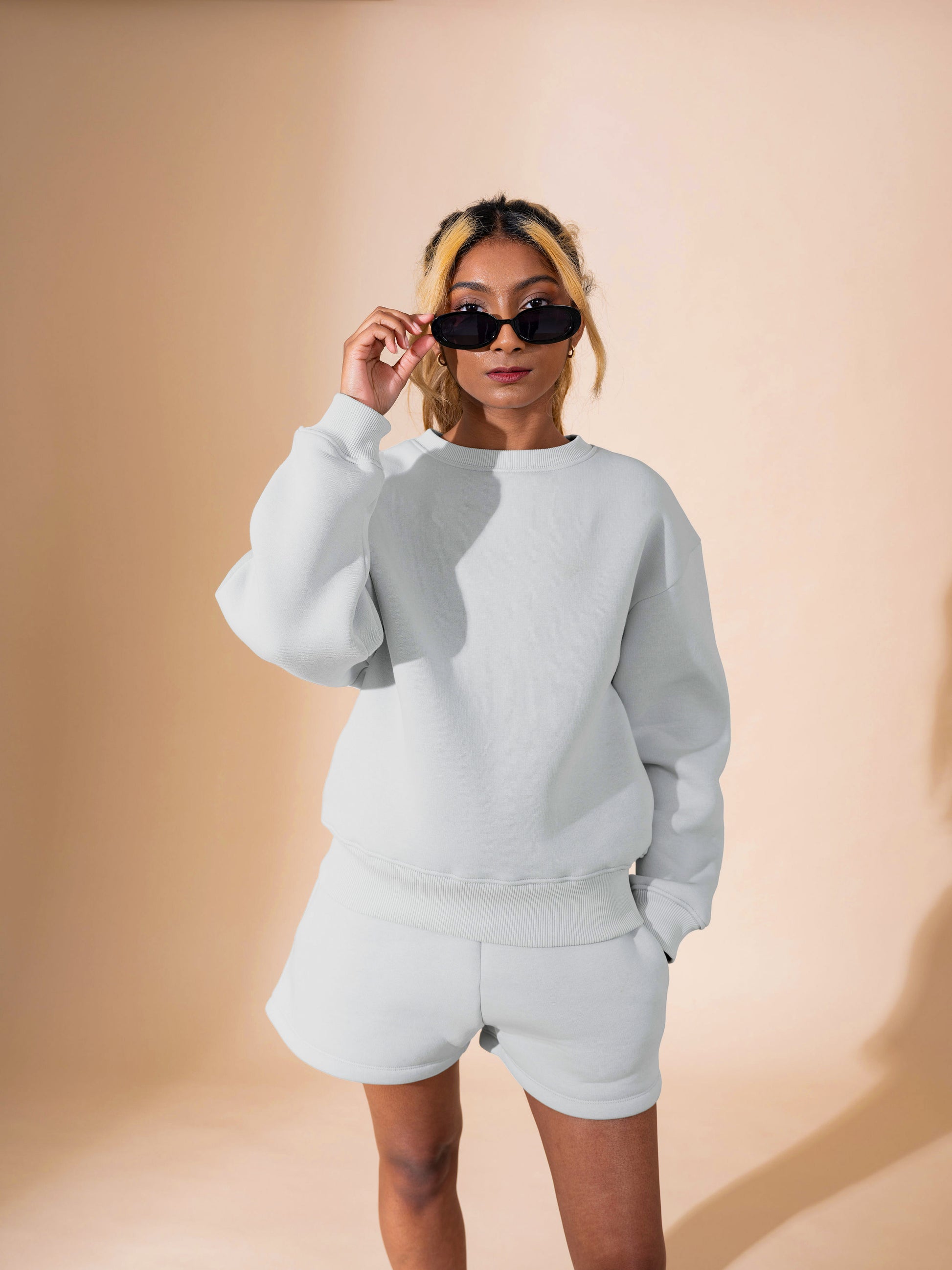 A person with light brown hair in a ponytail wears black sunglasses and the DEWEY BAILEY Crewneck Sweatshirt Jumper (Grey, Petite & Extra Petite), paired with matching shorts, standing against a beige background.