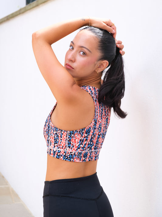 A woman in a DEWEY HAZEL Cropped Vest Top (Printed, Extra Petite) and black leggings stands sideways, looking at the camera. She lifts her dark ponytail against a white wall, showcasing bright athleisure style.