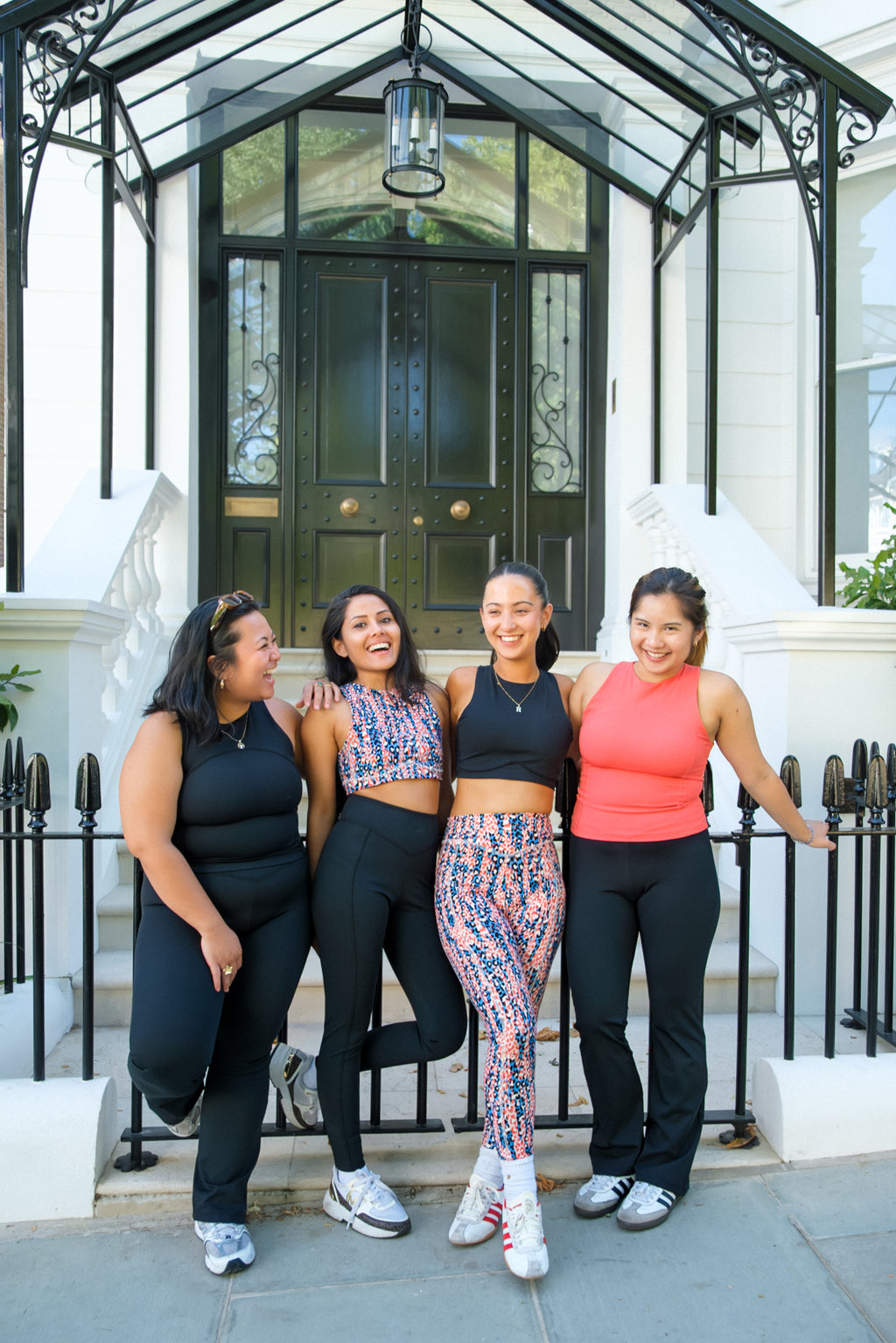 Four women in athleisure wear smile together outside a chic black door, radiating friendship after a workout. One wears the DEWEY HAZEL Cropped Vest Top (Black, Extra Petite), showcasing trendy petite activewear style.