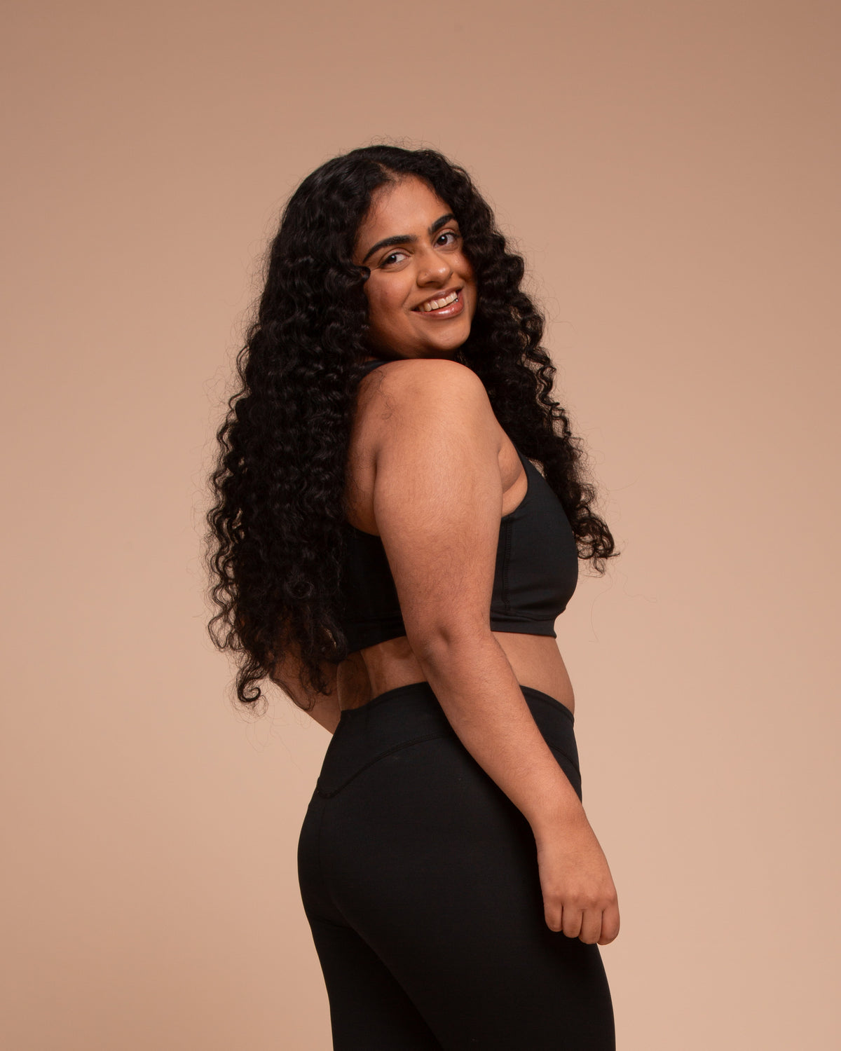 A smiling woman with long, curly hair models the DEWEY HAZEL Cropped Vest Top in black (Extra Petite), posing confidently against a beige background.