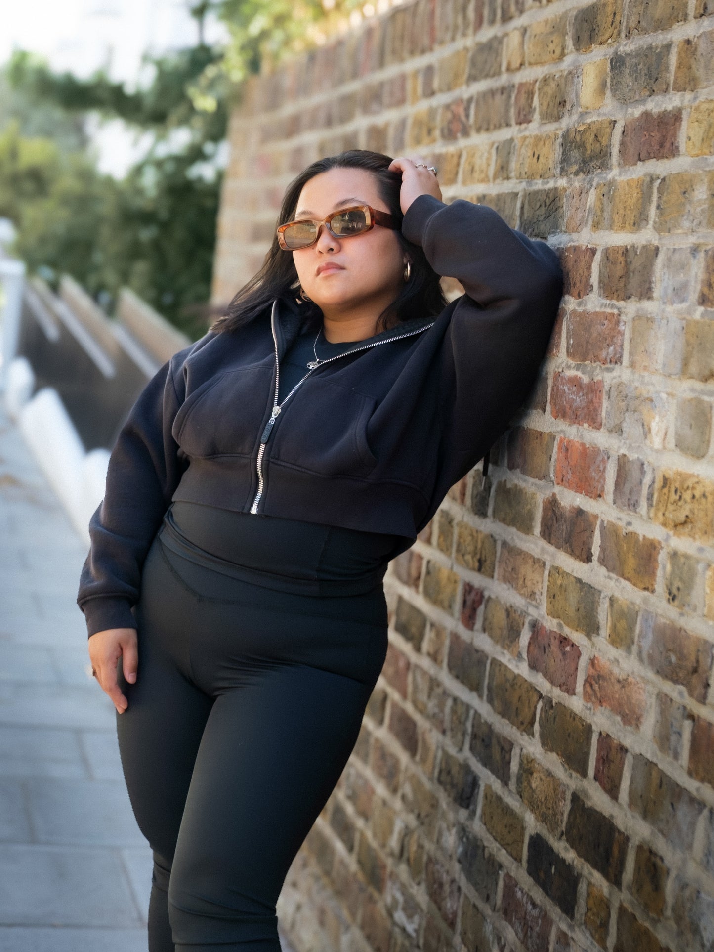 A woman in DEWEY’s TOBY Full Zip-Up Hoodie (Black, Extra Petite) and black leggings leans against a brick wall outdoors, confidently facing the camera with sunglasses on. Trees and a sidewalk are visible in the background.