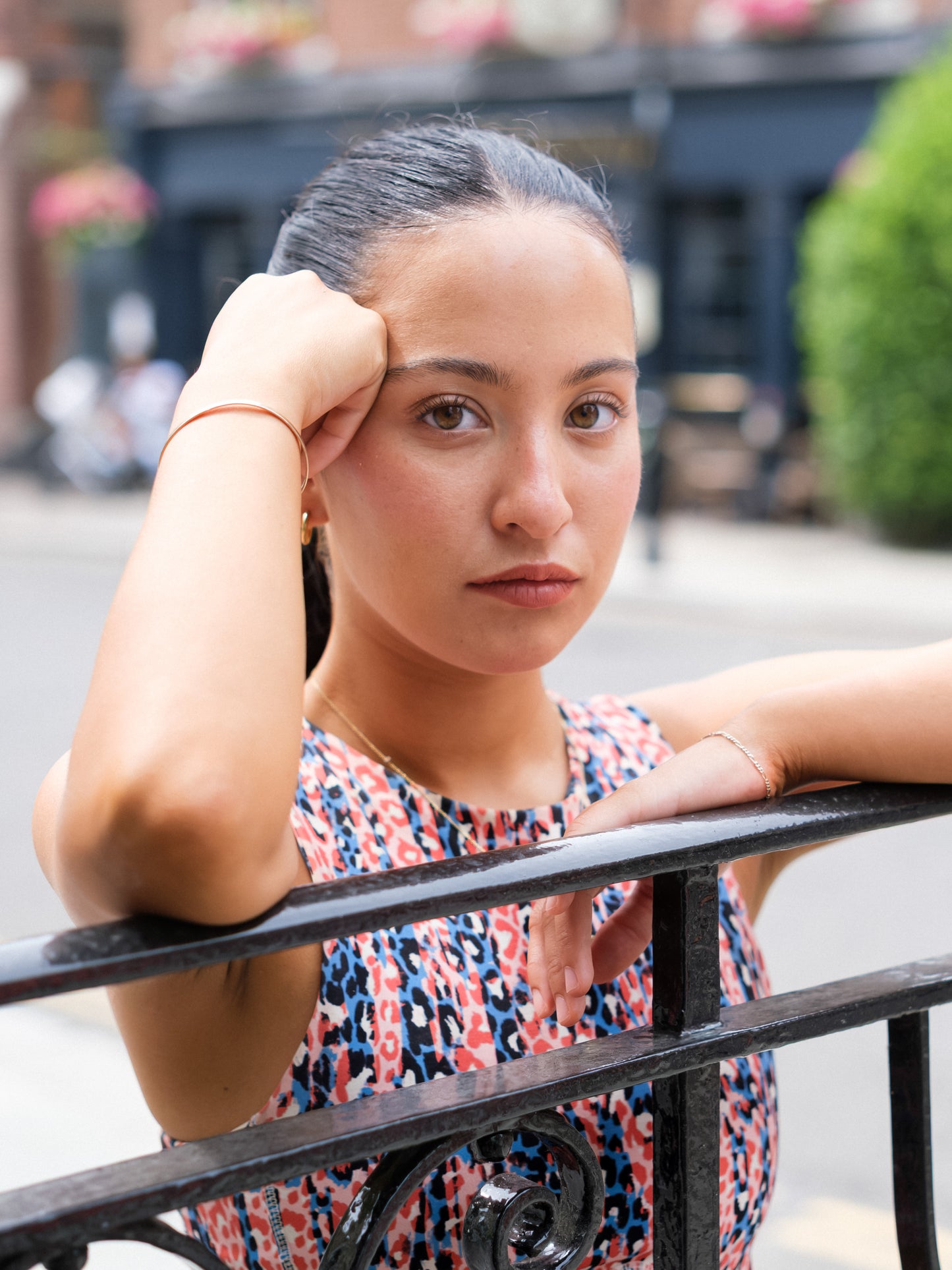 A woman with long dark hair pulled back, wearing a DEWEY HAZEL Cropped Vest Top (Printed, Extra Petite), leans on a black metal railing outdoors, resting her head on her hand and looking at the camera.