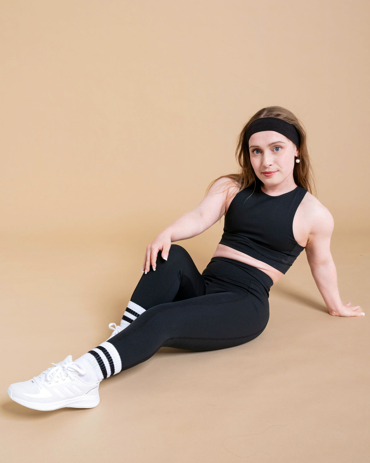 A woman wears the DEWEY HAZEL Cropped Vest Top in Black (Extra Petite) with white sneakers, sitting confidently on the floor against a beige background with one knee up and a hand behind her.