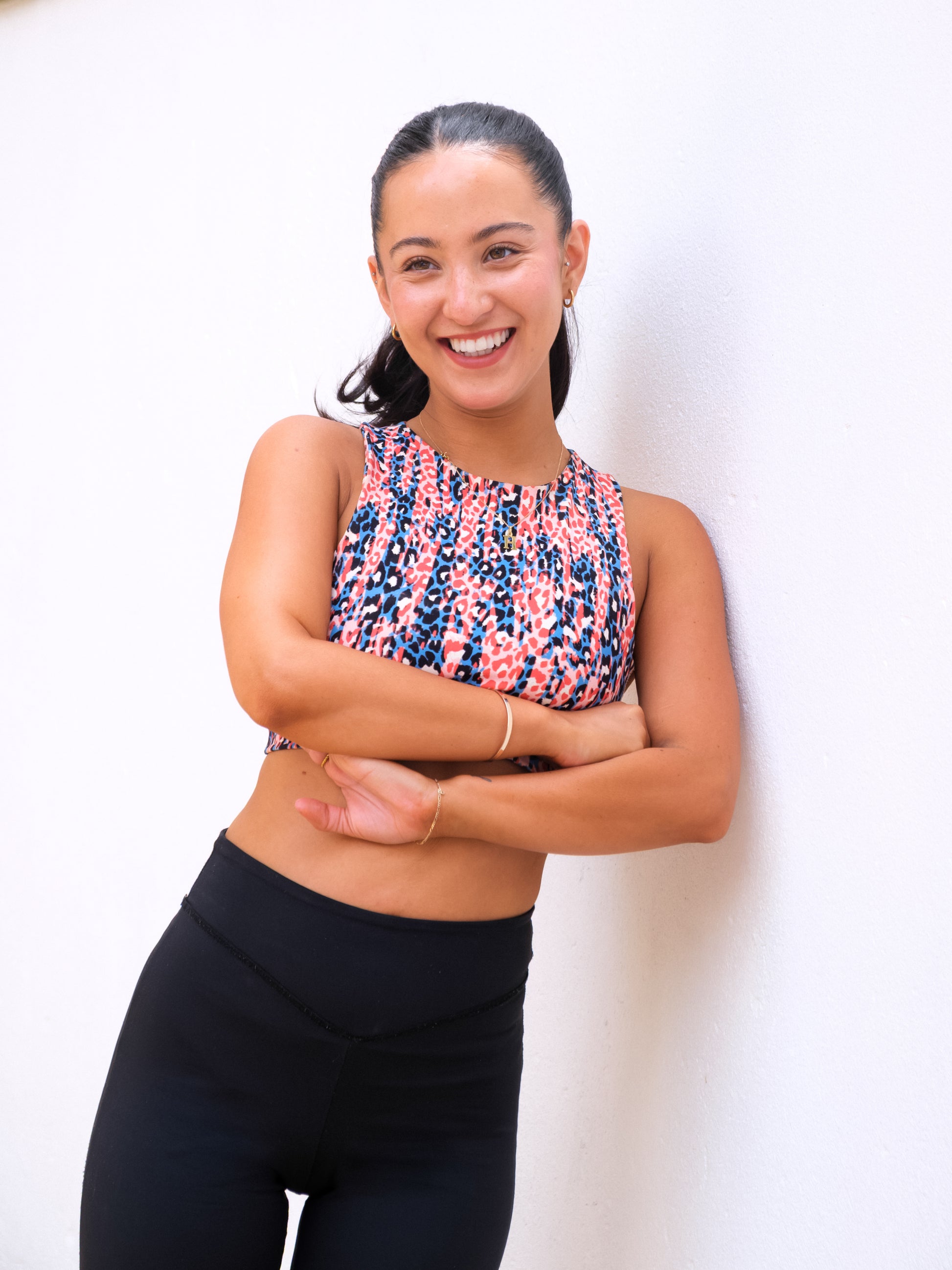 A young woman with long dark hair in a ponytail smiles, standing against a white wall with arms crossed. She wears the DEWEY HAZEL Cropped Vest Top (Printed, Extra Petite) and black leggings for a stylish athleisure look.