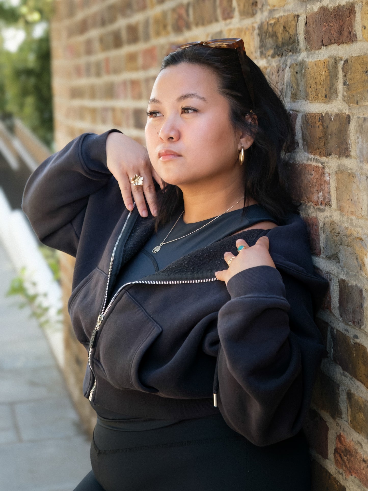 A woman with dark hair, wearing the DEWEY TOBY Full Zip-Up Hoodie in Black (Extra Petite), leans against a brick wall outdoors. She looks thoughtfully to the side, sunglasses on her head and one hand opening her jacket.