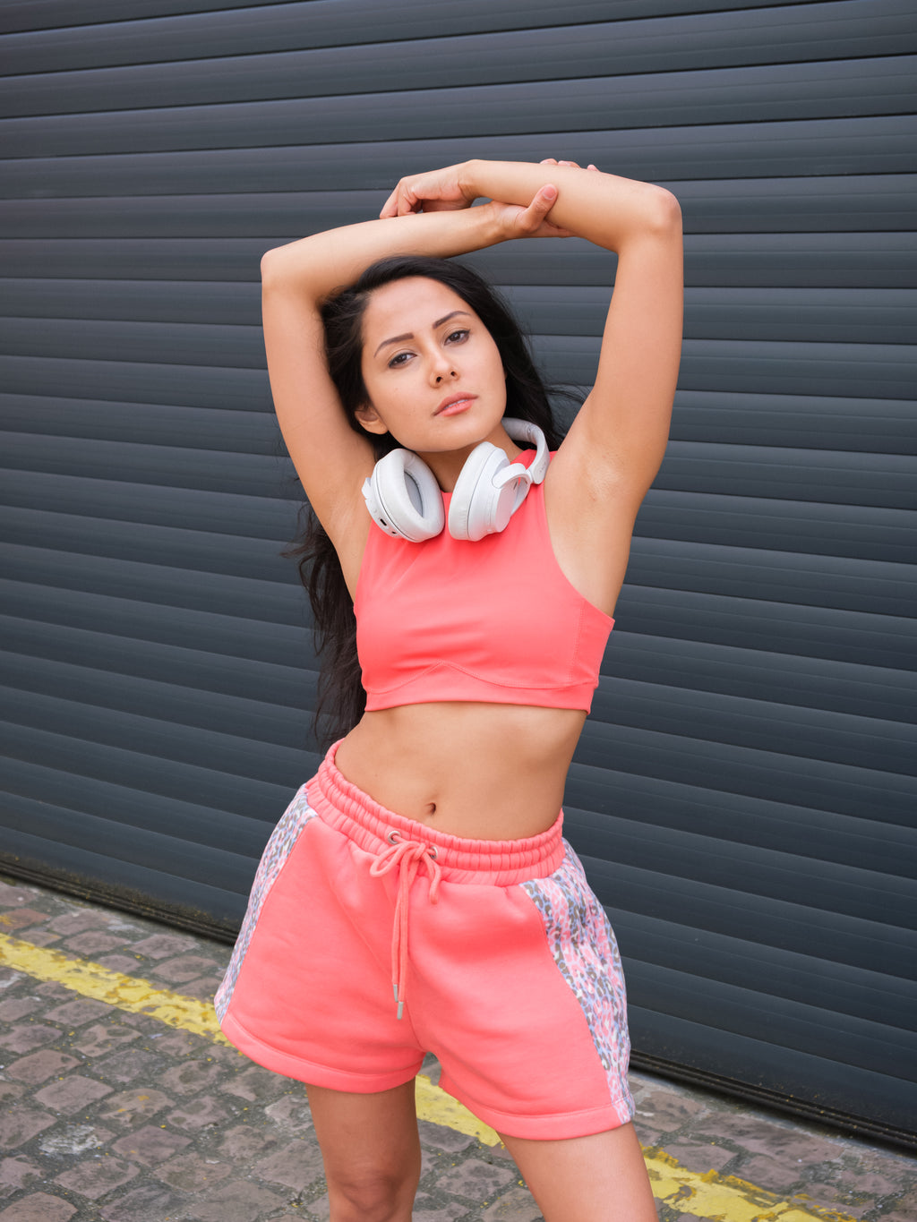 Wearing the DEWEY HAZEL Cropped Vest Top in Coral Red (Extra Petite), a woman with headphones around her neck stretches her arms above her head outdoors, standing before a dark corrugated metal wall.