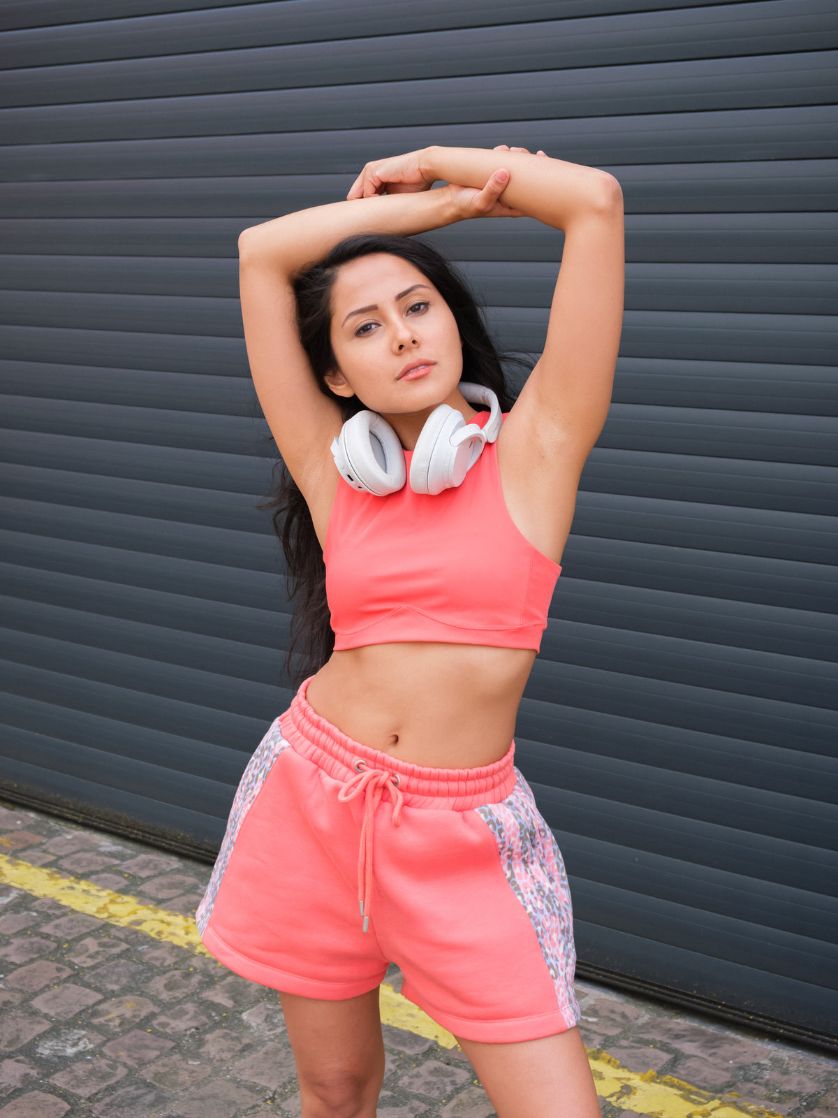 Wearing the DEWEY HAZEL Cropped Vest Top in Coral Red (Extra Petite), a woman with headphones around her neck stretches her arms above her head outdoors, standing before a dark corrugated metal wall.