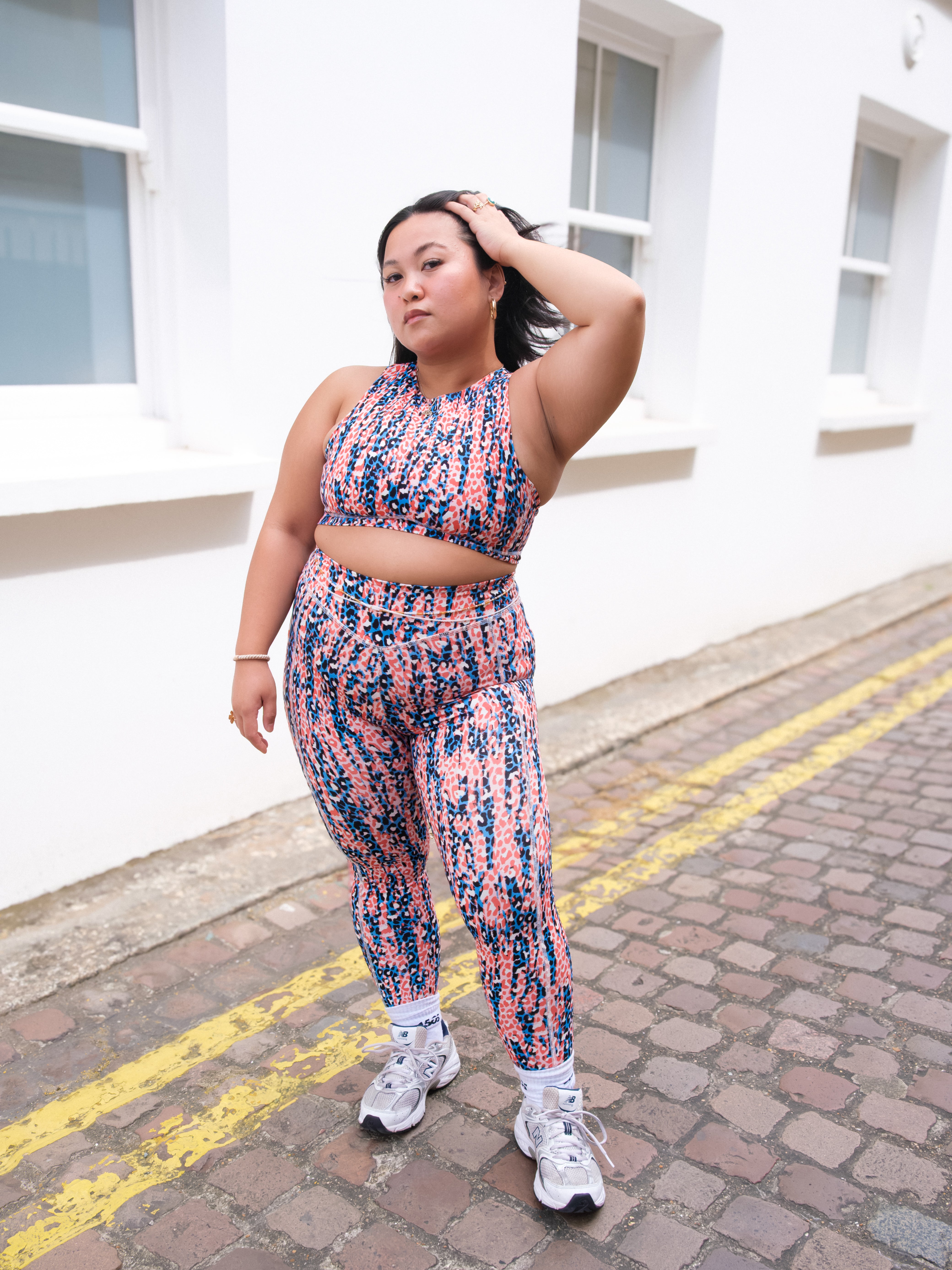 A woman poses on a cobblestone street by a white building, confidently wearing DEWEY’s IRIS High Waist Leggings (Printed, Extra Petite) with a matching sports bra and white sneakers, showcasing chic athleisure style.