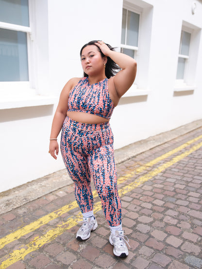 A woman poses on a cobblestone street by a white building, confidently wearing DEWEY’s IRIS High Waist Leggings (Printed, Extra Petite) with a matching sports bra and white sneakers, showcasing chic athleisure style.