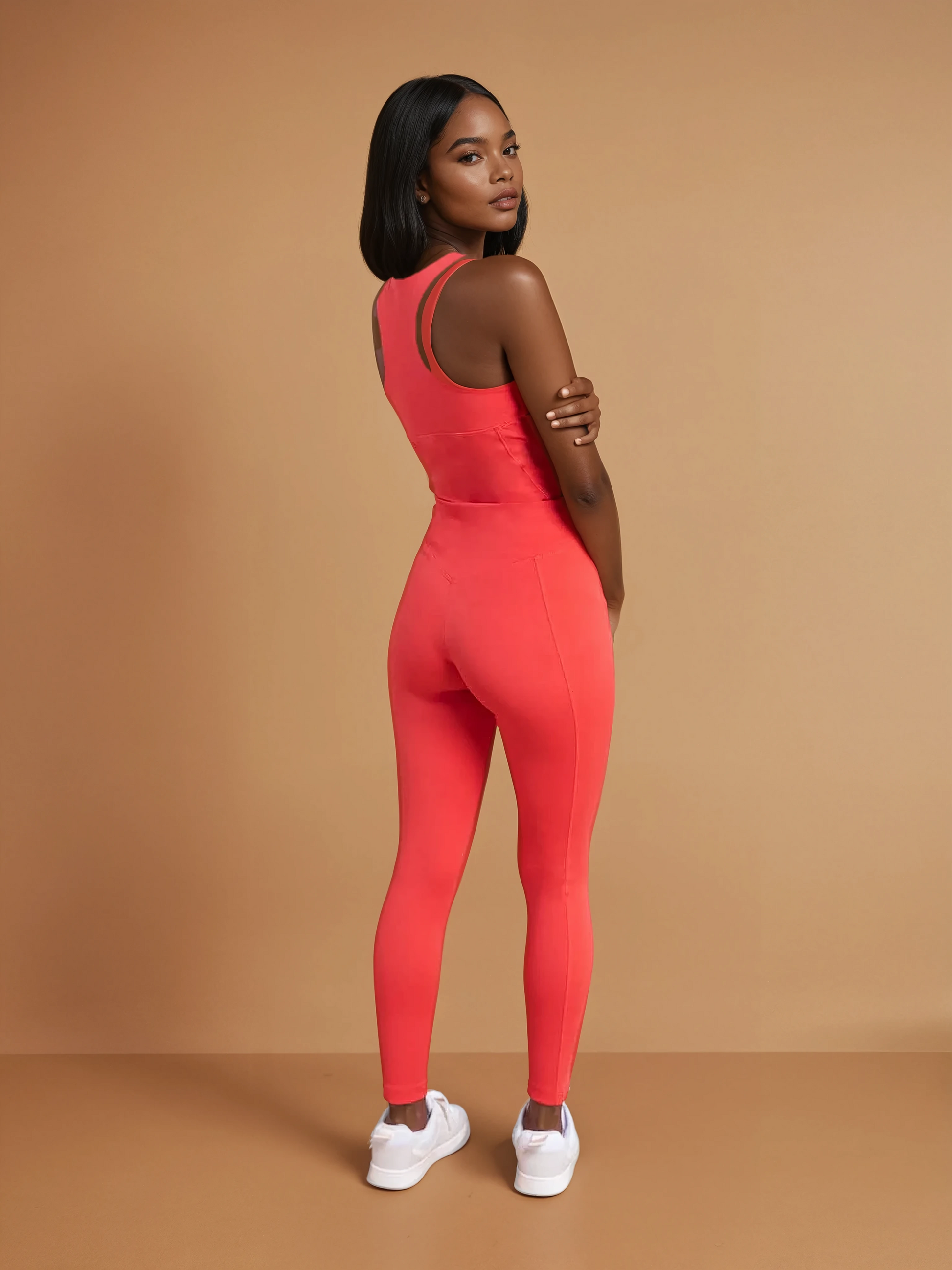 A woman stands with her back turned, looking over her shoulder. She wears DEWEY's IRIS High Waist Leggings in Coral Red (Extra Petite) with white sneakers, posed against a plain beige background.