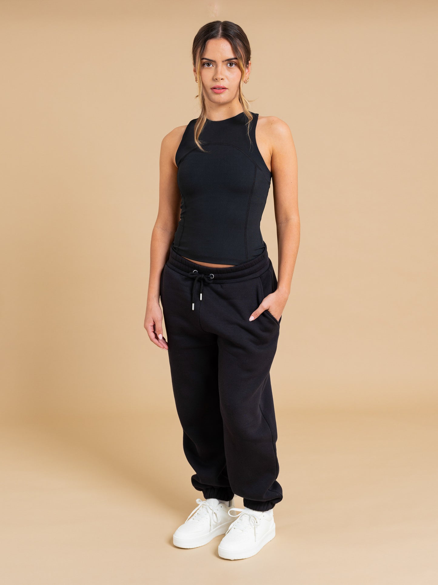 A woman stands against a beige background in the DEWEY SUMMER Racerback Vest Top (Black, Extra Petite), black drawstring sweatpants, and white sneakers, hands in pockets and looking directly at the camera.