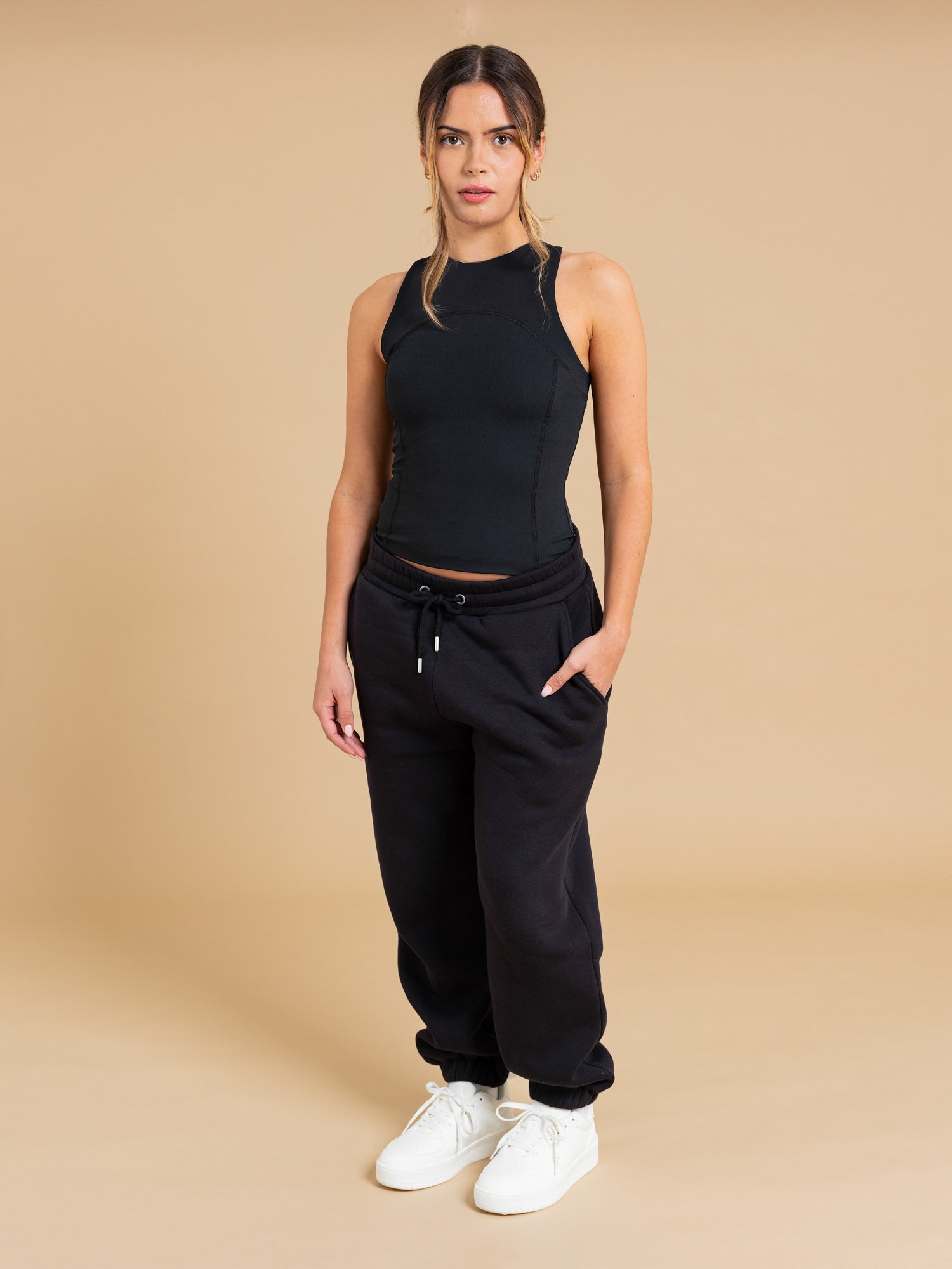 A woman stands against a beige background in the DEWEY SUMMER Racerback Vest Top (Black, Extra Petite), black drawstring sweatpants, and white sneakers, hands in pockets and looking directly at the camera.