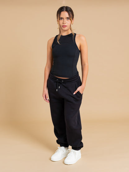 A woman stands against a beige background in the DEWEY SUMMER Racerback Vest Top (Black, Extra Petite), black drawstring sweatpants, and white sneakers, hands in pockets and looking directly at the camera.